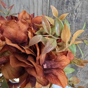 K&ouml;nnte beinhalten: Ein Strau&szlig; k&uuml;nstlicher Blumen mit Bl&uuml;tenbl&auml;ttern aus Stoff in tiefem Rostrot, gebranntem Orange und Braun. Das Arrangement enth&auml;lt schmale gr&uuml;ne und hellbraune Bl&auml;tter an d&uuml;nnen Stielen sowie ein kleines rosa Detail in der Mitte einer Bl&uuml;te.