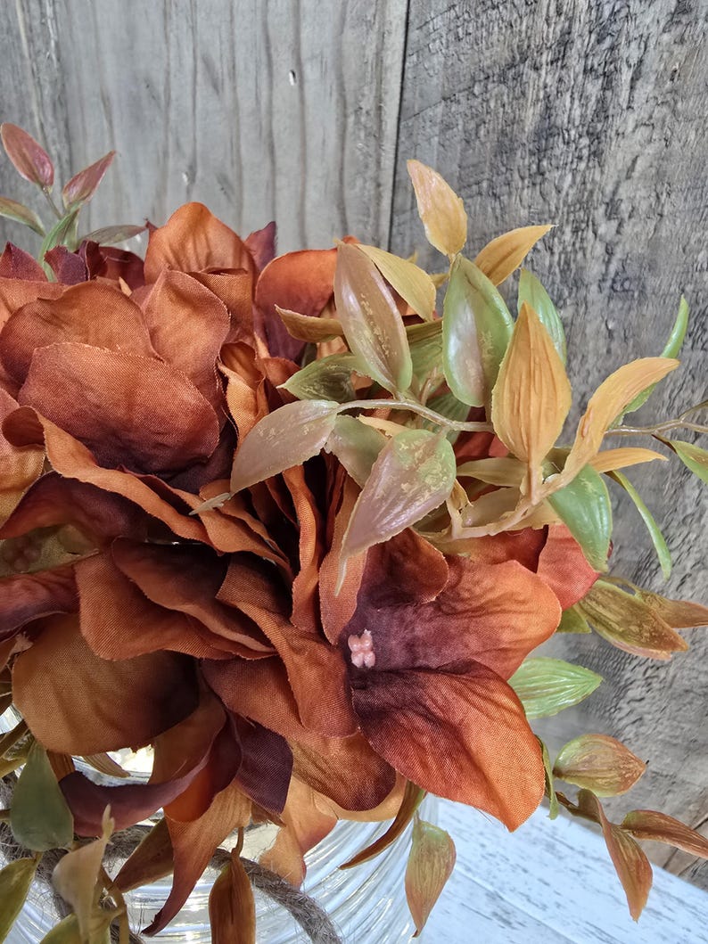 K&ouml;nnte beinhalten: Ein Strau&szlig; k&uuml;nstlicher Blumen mit Bl&uuml;tenbl&auml;ttern aus Stoff in tiefem Rostrot, gebranntem Orange und Braun. Das Arrangement enth&auml;lt schmale gr&uuml;ne und hellbraune Bl&auml;tter an d&uuml;nnen Stielen sowie ein kleines rosa Detail in der Mitte einer Bl&uuml;te.