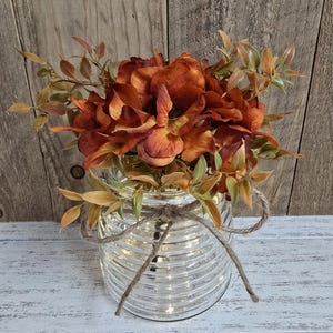 K&ouml;nnte beinhalten: Ein klares, horizontal geripptes Glasgef&auml;&szlig;, das mit einer braunen Schnur um den Hals gebunden ist. Das Gef&auml;&szlig; ist mit k&uuml;nstlichen Blumen in den Farben gebranntes Orange und tiefes Kupfer gef&uuml;llt, akzentuiert durch kleine hellgr&uuml;ne und beige Blattstiele.
