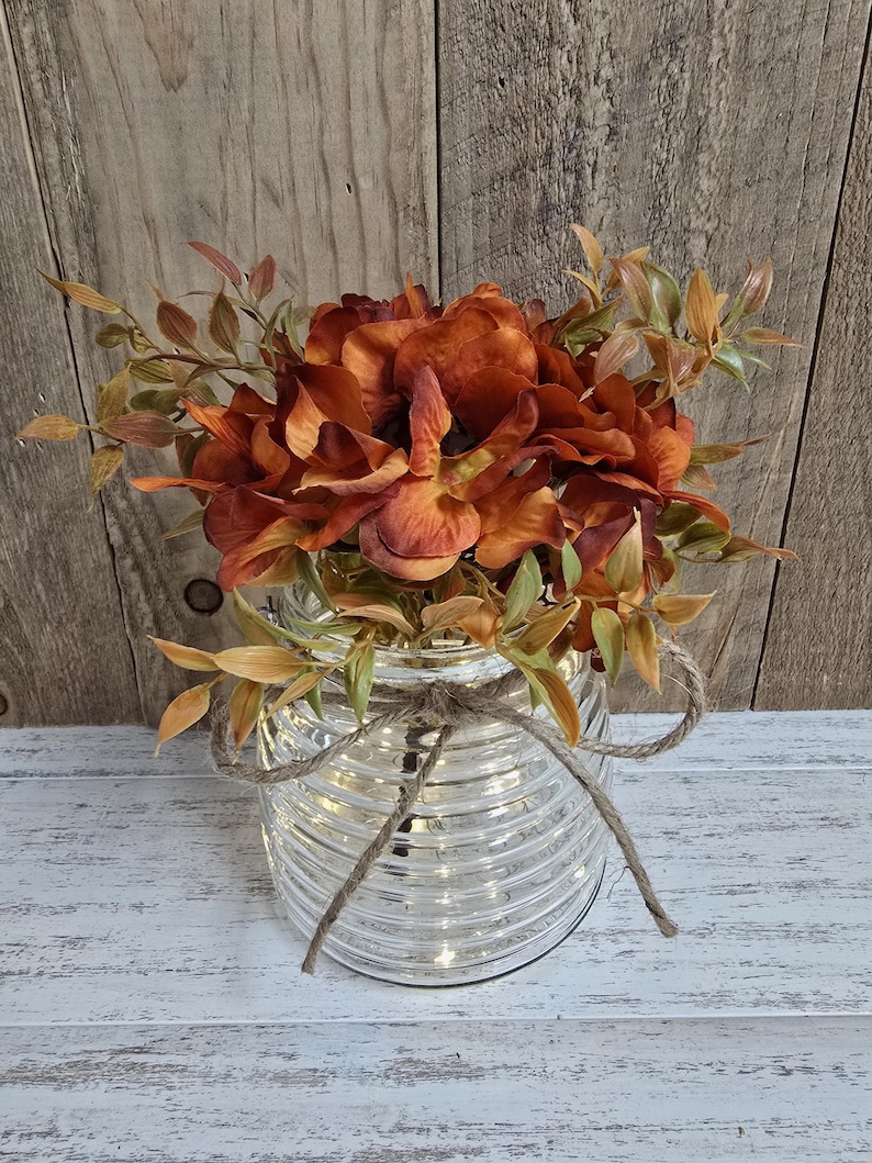 K&ouml;nnte beinhalten: Ein klares, horizontal geripptes Glasgef&auml;&szlig;, das mit einer braunen Schnur um den Hals gebunden ist. Das Gef&auml;&szlig; ist mit k&uuml;nstlichen Blumen in den Farben gebranntes Orange und tiefes Kupfer gef&uuml;llt, akzentuiert durch kleine hellgr&uuml;ne und beige Blattstiele.