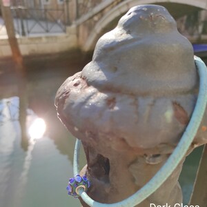 May include: A blue glass bead with a silver charm on a light blue leather bracelet. The bracelet is wrapped around a dark gray metal post. The text "Dark Glass" is visible in the image.