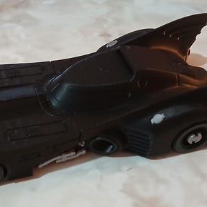 Op de afbeelding: Een zwart speelgoedauto dat lijkt op de Batmobile, met een gestroomlijnd ontwerp en opvallende vinnen. De auto heeft gedetailleerde kenmerken, waaronder wielen en een cockpit. Het is waarschijnlijk een verzamelobject.