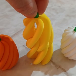 Puede incluir: Un surtido de calabazas decorativas con forma de calabaza, un racimo de plátanos y una calabaza blanca. La calabaza es naranja, los plátanos son amarillos y la calabaza blanca tiene un tallo verde. Los objetos son de un material liso.