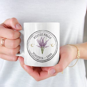 May include: White ceramic mug featuring a circular design with the text "Mystic Falls Virginia - 1864". The design includes a bouquet of purple flowers, crescent moons, and stars. The mug is held by a person.