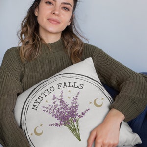 May include: Cream-colored throw pillow with a circular design featuring the text "MYSTIC FALLS VIRGINIA" and a purple lavender bouquet. The design includes gold crescent moons and stars. The pillow is held by a person wearing a green sweater.