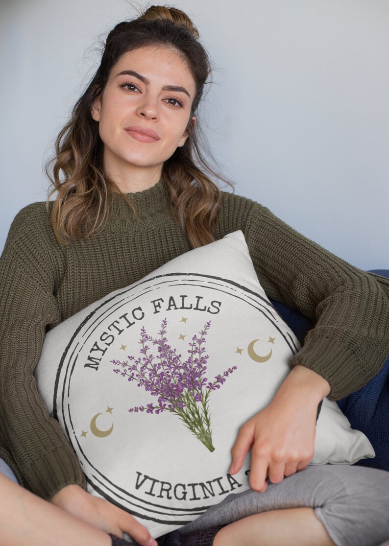 May include: Cream-colored throw pillow with a circular design featuring the text "MYSTIC FALLS VIRGINIA" and a purple lavender bouquet. The design includes gold crescent moons and stars. The pillow is held by a person wearing a green sweater.