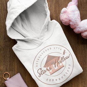 May include: White hooded sweatshirt with a hood, displaying a circular design with the text "Stars Hollow Connecticut" and a building illustration. A pink key card holder and a pink plush toy are also visible.