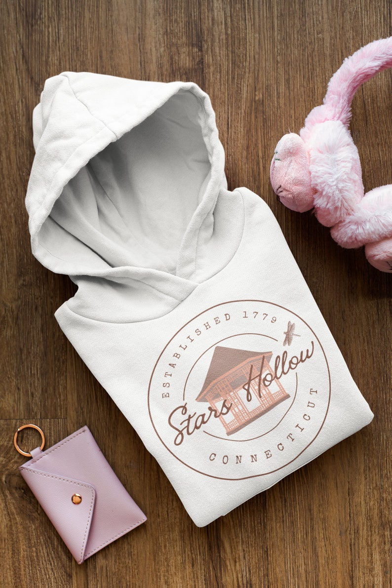 May include: White hooded sweatshirt with a hood, displaying a circular design with the text "Stars Hollow Connecticut" and a building illustration. A pink key card holder and a pink plush toy are also visible.