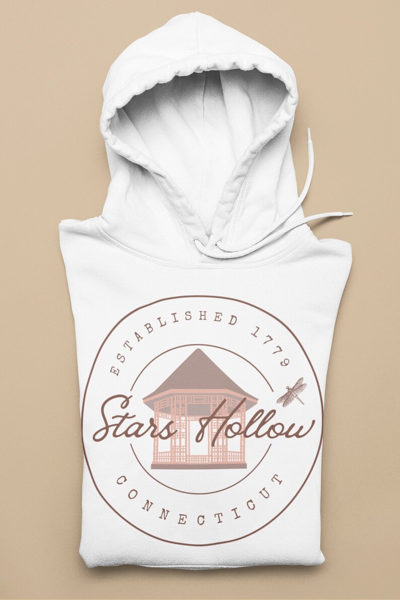 May include: White hoodie with a drawstring. The front displays a circular design with the words "Stars Hollow Connecticut" and a gazebo illustration. The design also includes the text "ESTABLISHED 1779" and a dragonfly.