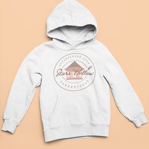 May include: White hoodie with a front pocket and a drawstring hood. The sweatshirt displays a circular graphic with the text "ESTABLISHED 1779 Stars Hollow CONNECTICUT" around a gazebo illustration and a dragonfly.