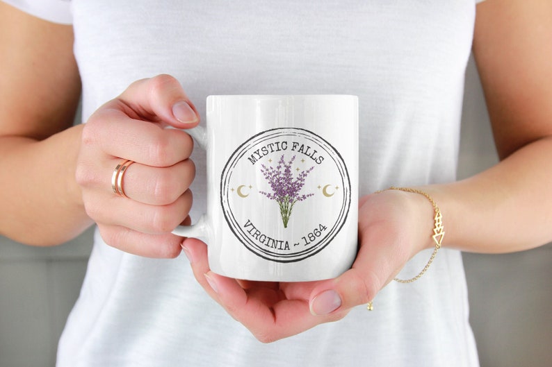 May include: White ceramic mug featuring a circular design with the text "Mystic Falls Virginia - 1864". The design includes a bouquet of purple flowers, crescent moons, and stars. The mug is held by a person.