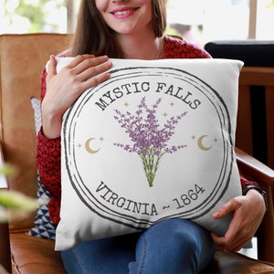 May include: White square pillow with a circular design featuring the text "Mystic Falls Virginia ~ 1864". The design includes a bouquet of purple flowers, gold crescent moons, and stars.