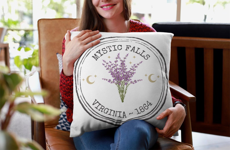 May include: White square pillow with a circular design featuring the text "Mystic Falls Virginia ~ 1864". The design includes a bouquet of purple flowers, gold crescent moons, and stars.