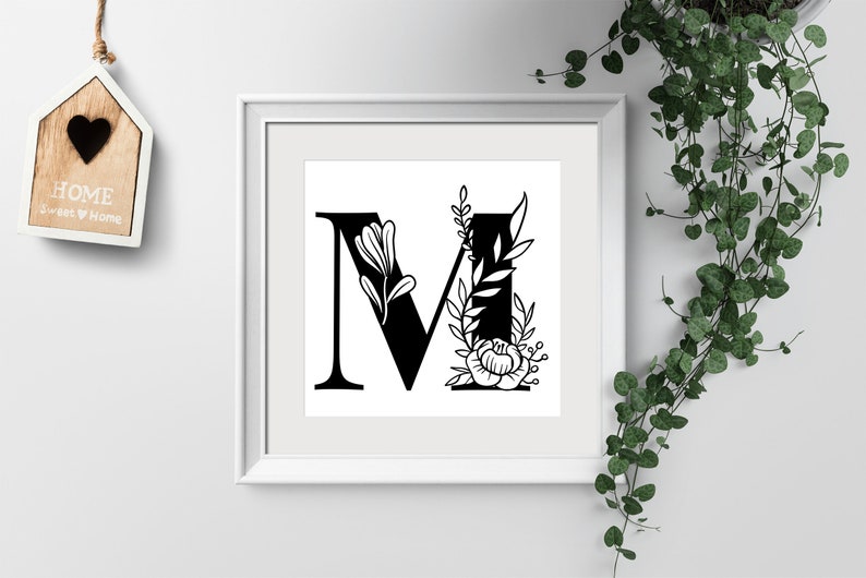 May include: A black and white printable art print featuring the letter M with floral embellishments. The letter is in a bold, serif font and is surrounded by stylized leaves and flowers.