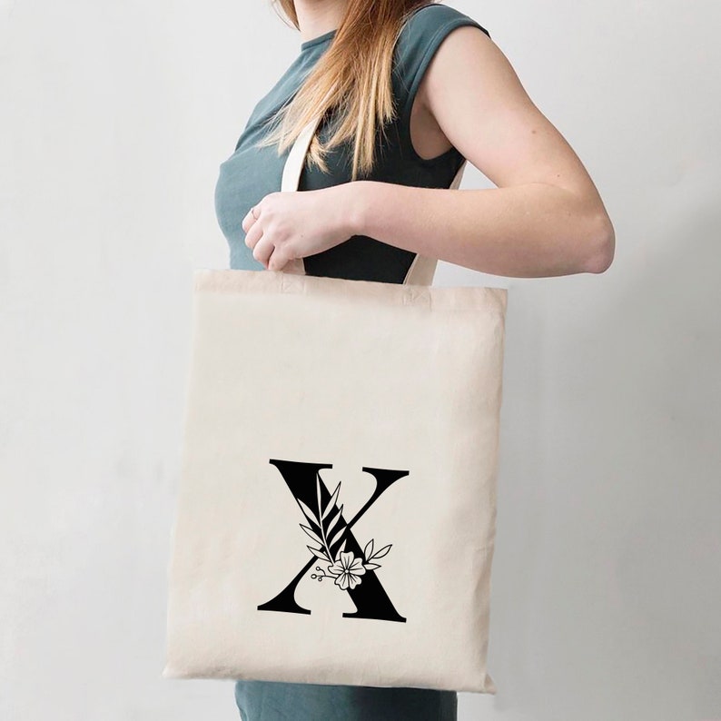May include: A white canvas tote bag with a black floral design featuring the letter X. The bag has black handles.