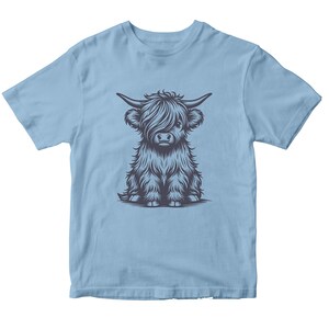 May include: A light blue t-shirt with a graphic of a cute, cartoon Highland cow with black fur. The cow is sitting down with its legs crossed.
