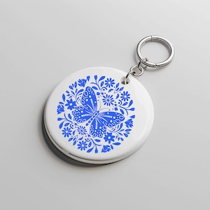 May include: A white ceramic keychain with a blue butterfly design surrounded by flowers. The keychain is attached to a silver metal ring.