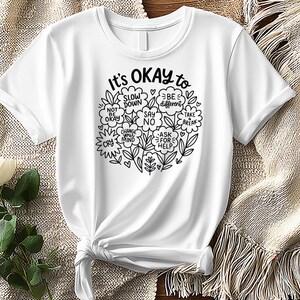 Op de afbeelding: Wit t-shirt met een zwarte inkt ronde afbeelding met de tekst "It's OKAY to" met zinnen als "Slow Down" en "Ask for Help" in bloemen- en bladmotieven. Het t-shirt is aan de onderkant geknoopt.