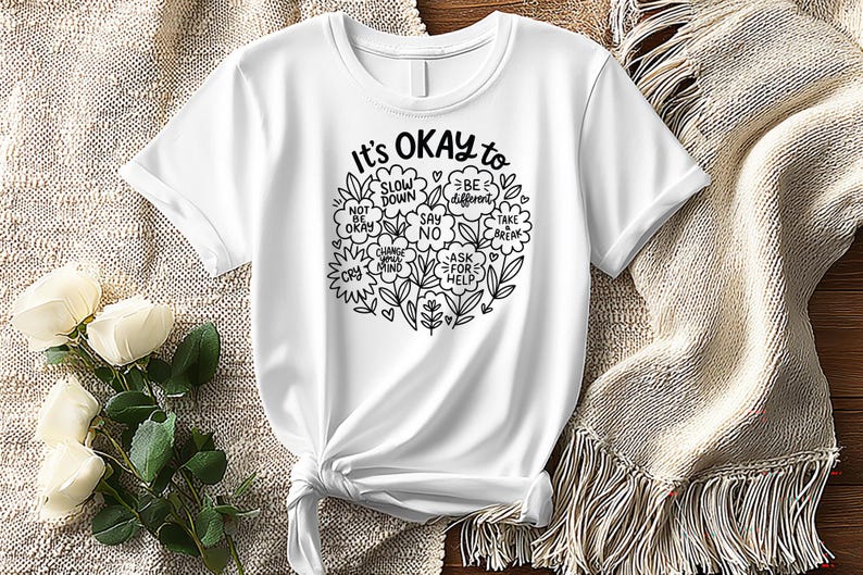 Op de afbeelding: Wit t-shirt met een zwarte inkt ronde afbeelding met de tekst "It's OKAY to" met zinnen als "Slow Down" en "Ask for Help" in bloemen- en bladmotieven. Het t-shirt is aan de onderkant geknoopt.