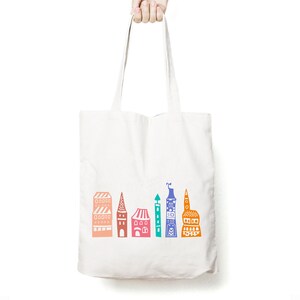May include: White canvas tote bag with a colorful illustration of a city skyline. The buildings are in shades of orange, pink, blue, green, and purple.