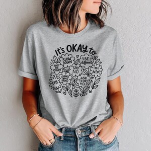 Op de afbeelding: Een lichtgrijs t-shirt met de tekst "It's OKAY to" in zwart schrift, omringd door een bloemenontwerp. De boodschap van het shirt bevat zinnen als "Rustig aan" en "Anders zijn".