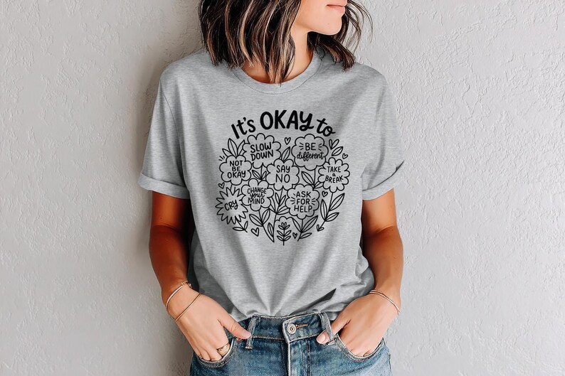 Op de afbeelding: Een lichtgrijs t-shirt met de tekst "It's OKAY to" in zwart schrift, omringd door een bloemenontwerp. De boodschap van het shirt bevat zinnen als "Rustig aan" en "Anders zijn".