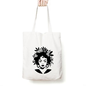 May include: A white tote bag with a black silhouette of a woman with flowers in her hair. The woman is wearing large earrings.