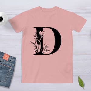 May include: A pink t-shirt with a black letter D and floral design. The letter D is outlined in black and filled in with black. The floral design is black and features a tulip and leaves.