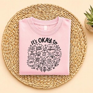 Op de afbeelding: Een lichtroze sweatshirt met de tekst "It's OKAY to" omringd door bloemen en motiverende woorden. De tekst bevat zinnen als "Slow Down", "Be Different" en "Ask for Help". Het sweatshirt ligt op een geweven placemat, met een kleine potplant.