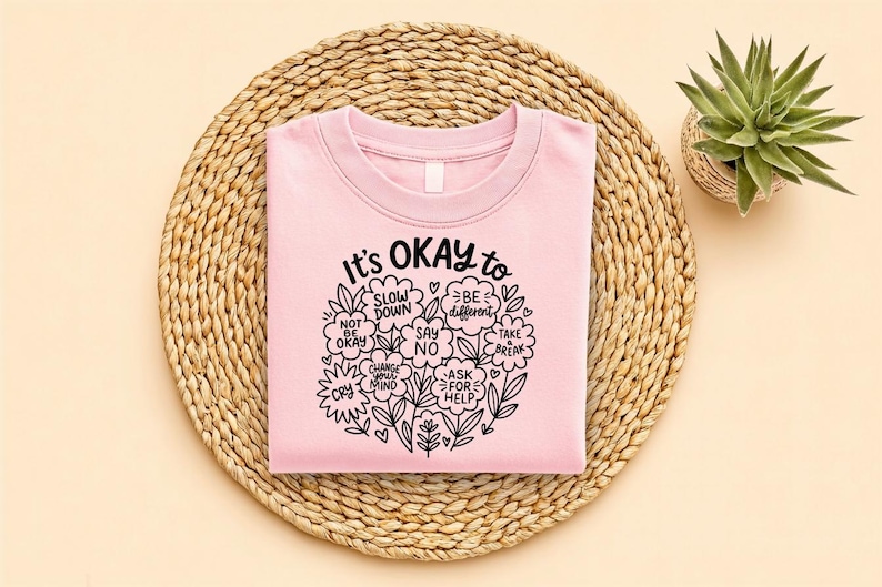 Op de afbeelding: Een lichtroze sweatshirt met de tekst "It's OKAY to" omringd door bloemen en motiverende woorden. De tekst bevat zinnen als "Slow Down", "Be Different" en "Ask for Help". Het sweatshirt ligt op een geweven placemat, met een kleine potplant.