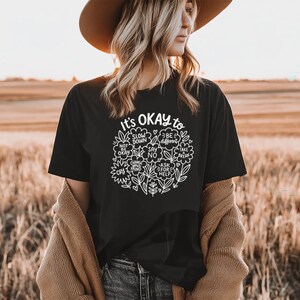 Op de afbeelding: Zwart t-shirt met de tekst "It's OKAY to" en een bloemenontwerp. Het ontwerp bevat zinnen als "Slow Down", "Be Different" en "Ask for Help". Het shirt wordt gedragen met een spijkerbroek en een bruine hoed.