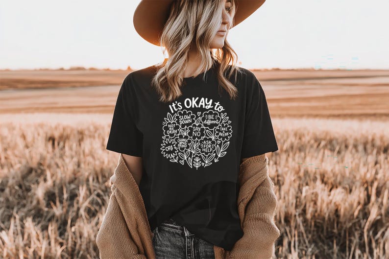 Op de afbeelding: Zwart t-shirt met de tekst "It's OKAY to" en een bloemenontwerp. Het ontwerp bevat zinnen als "Slow Down", "Be Different" en "Ask for Help". Het shirt wordt gedragen met een spijkerbroek en een bruine hoed.