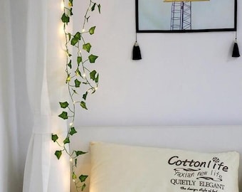 May include: A string of fairy lights wrapped around a faux ivy garland. The garland is hanging on a white wall next to a white bed with a cream pillow. The pillow has the text "Cottonlife Yaxian new life QUIETLY ELEGANT the design style of led HOME" printed on it.