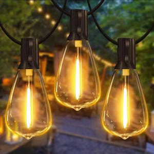 May include: String of three clear, teardrop-shaped light bulbs with a warm, yellow glow. Each bulb has a black base and is suspended from a black wire. The background is blurred, suggesting an outdoor setting, possibly a patio or garden.