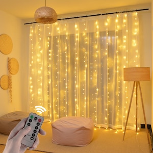 May include: A set of warm white LED fairy lights strung on a sheer white curtain. The lights are controlled by a remote with various settings. The lights are hanging in a room with a beige wall and a wooden floor.