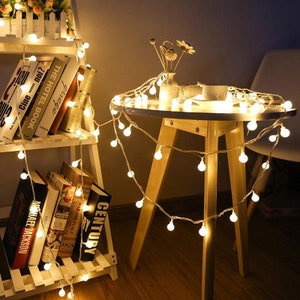 May include: A string of warm white LED fairy lights with round bulbs wrapped around a wooden table and a white bookshelf. The lights are strung around a vase with a flower arrangement and a white cup.