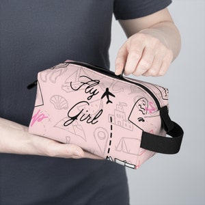 May include: Pink travel-themed cosmetic bag with a black zipper and handle. The bag has a white and black graphic print with the text "Fly Girl" and various travel icons.