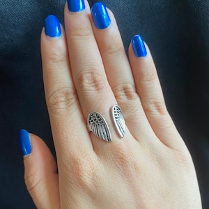 May include: A silver ring with two angel wings on the band. The ring is on a finger with blue nail polish.