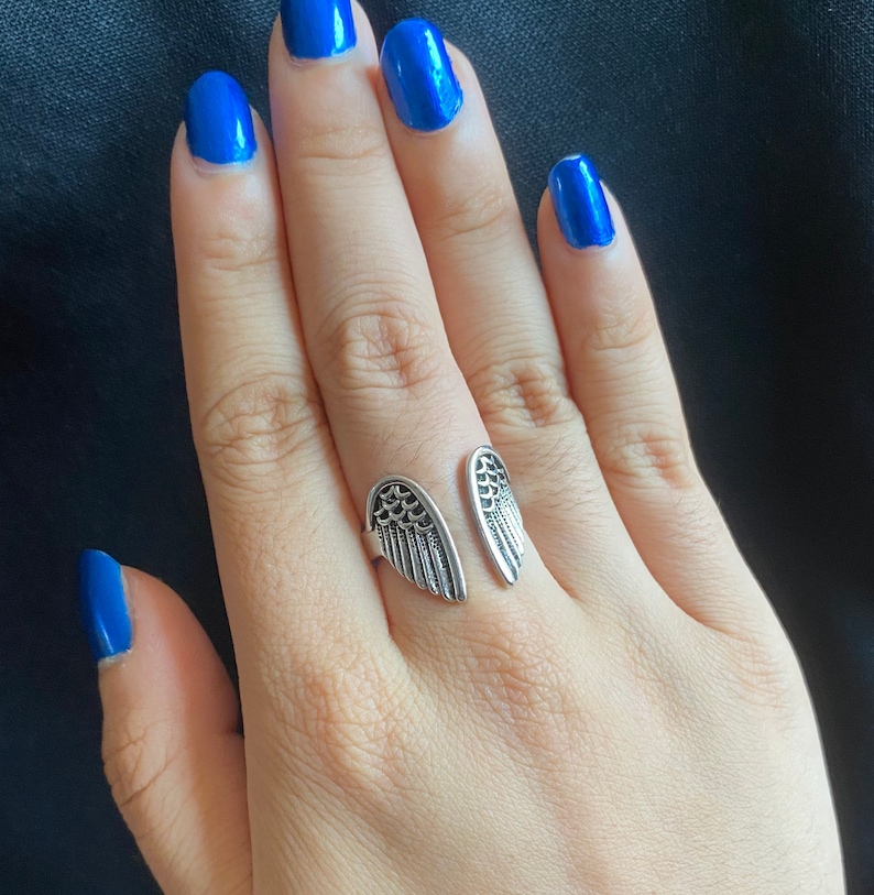 May include: A silver ring with two angel wings on the band. The ring is on a finger with blue nail polish.
