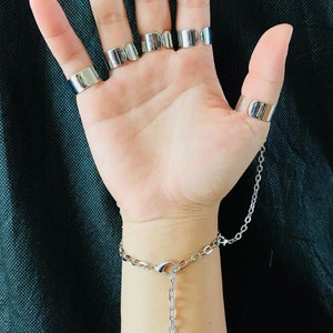 May include: A silver chain bracelet with a dangling sword charm. The bracelet has a clasp closure and is connected to a series of silver rings worn on the fingers.