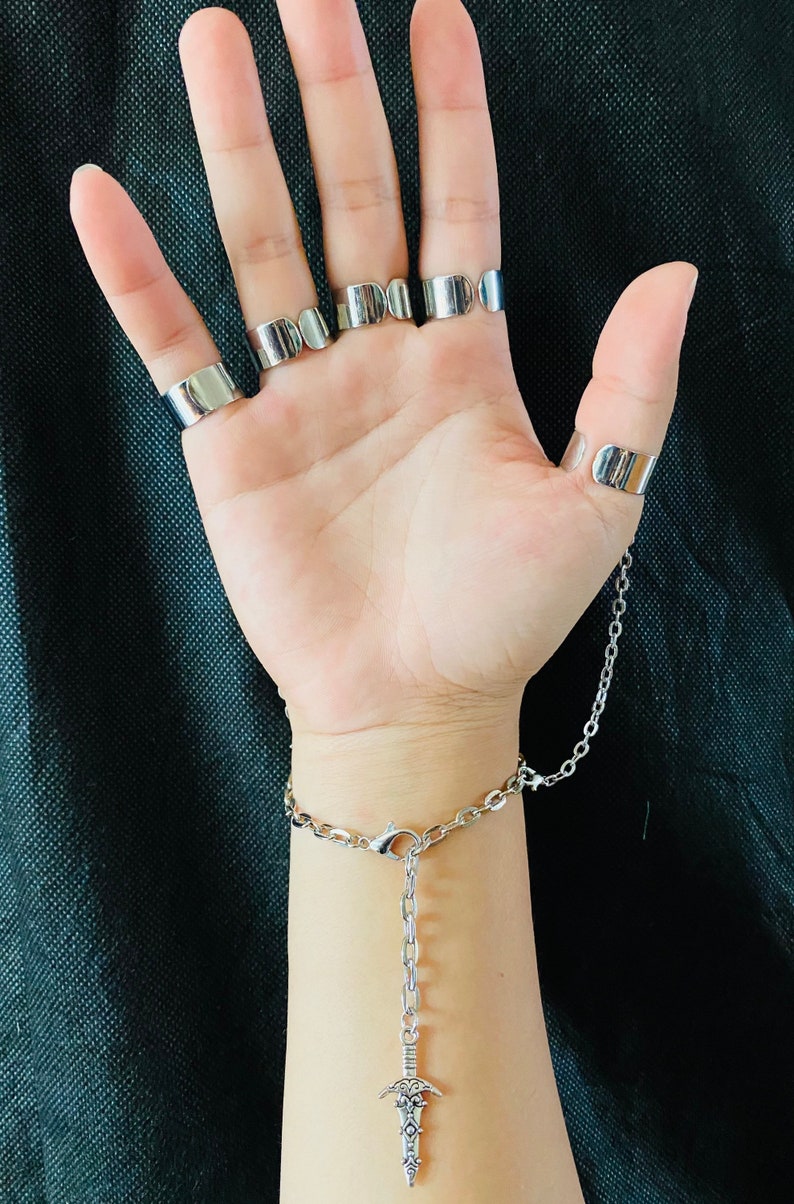 May include: A silver chain bracelet with a dangling sword charm. The bracelet has a clasp closure and is connected to a series of silver rings worn on the fingers.