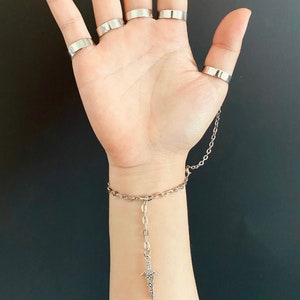K&ouml;nnte beinhalten: Ein silbernes Kettenarmband mit einem baumelnden Dolch-Anh&auml;nger. Das Armband hat mehrere silberne Ringe daran befestigt.