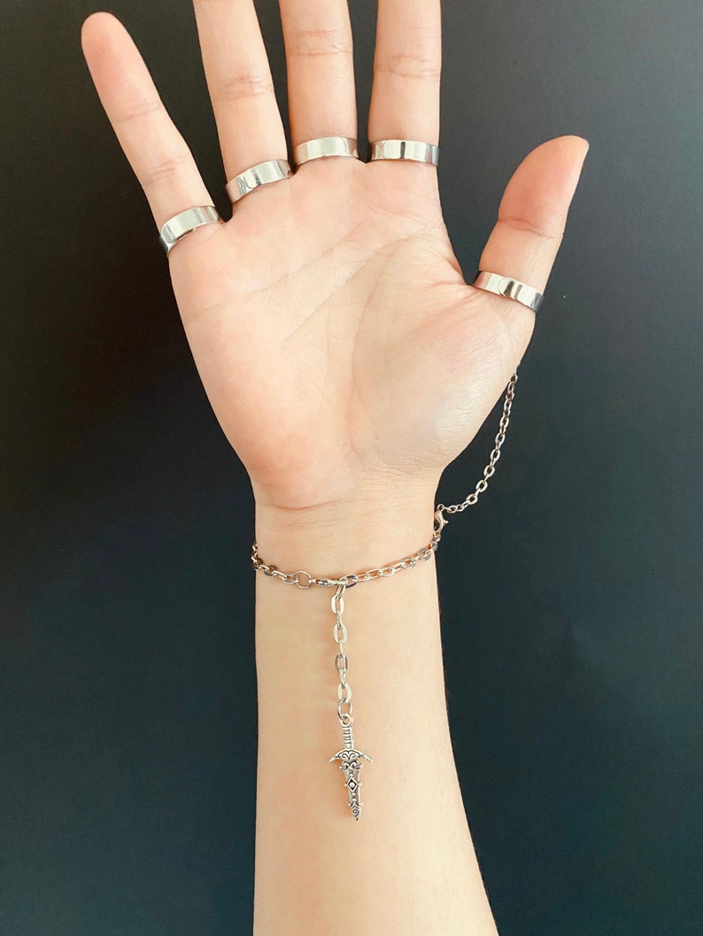 K&ouml;nnte beinhalten: Ein silbernes Kettenarmband mit einem baumelnden Dolch-Anh&auml;nger. Das Armband hat mehrere silberne Ringe daran befestigt.