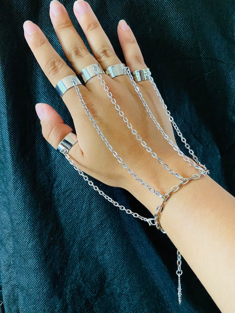 May include: A set of five silver rings connected by chains. The rings are worn on the fingers of one hand, and the chains extend down to the wrist, forming a bracelet.