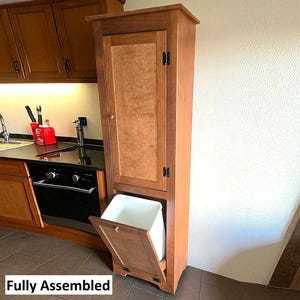 May include: A tall, wooden kitchen cabinet with a pull-down door revealing a white trash bin. The cabinet is a warm brown color and has a single door on top. The text "Fully Assembled" is at the bottom.