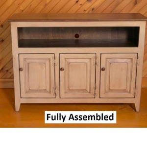 May include: A white wooden entertainment center with three doors and a shelf. The doors have raised panels and the top is a darker wood. The text "Fully Assembled" is printed below the entertainment center.
