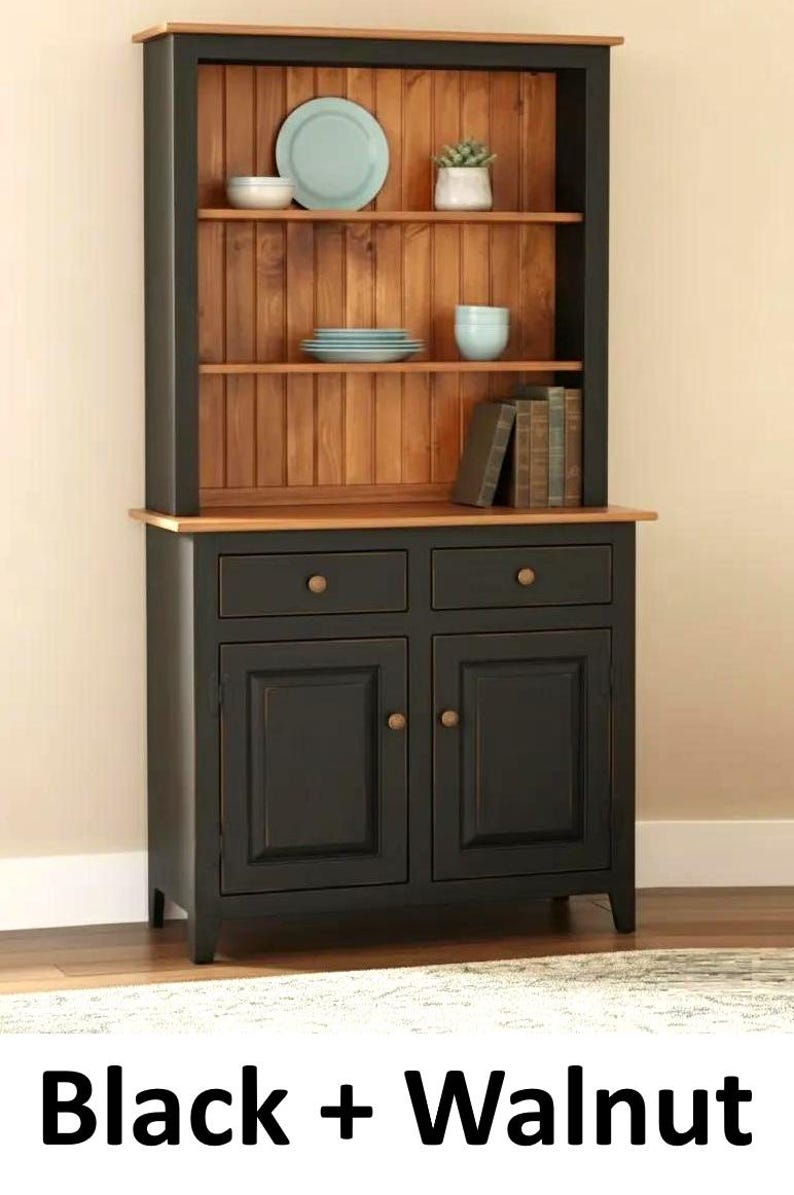 May include: A black and walnut hutch featuring two lower cabinet doors, two drawers with round knobs, and two open shelves with a vertical plank back. The unit displays plates, bowls, and books. The text Black + Walnut is visible below.