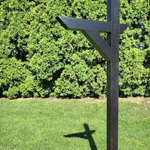 May include: A black wooden mailbox post with a horizontal arm extending out. The post is standing in a grassy area with a green hedge in the background.