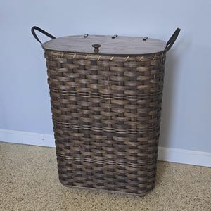 May include: A tall, rectangular brown woven laundry hamper with a wooden lid. The hamper has two black handles and a wooden knob on the lid. The hamper is made of woven strips of brown material.