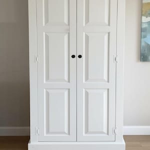 以下が含まれることがあります： 凹型パネル、黒い円形のノブ、黒い天板を備えた2枚扉の背の高い白いキャビネット。扉の外側には白い蝶番が見えます。画像には「Pure White + Black (Base) (Countertop)」というテキストが含まれています。